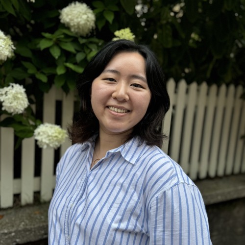 head and shoulders portrait of adult with shoulder length dark hair wearing a blue and white striped shirt in front of white hydrangeas and a picket fence